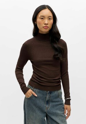 Femme aux longs cheveux foncés porte un pull à col roulé marron et un jean bleu taille haute, debout avec une main dans la poche contre un fond blanc.