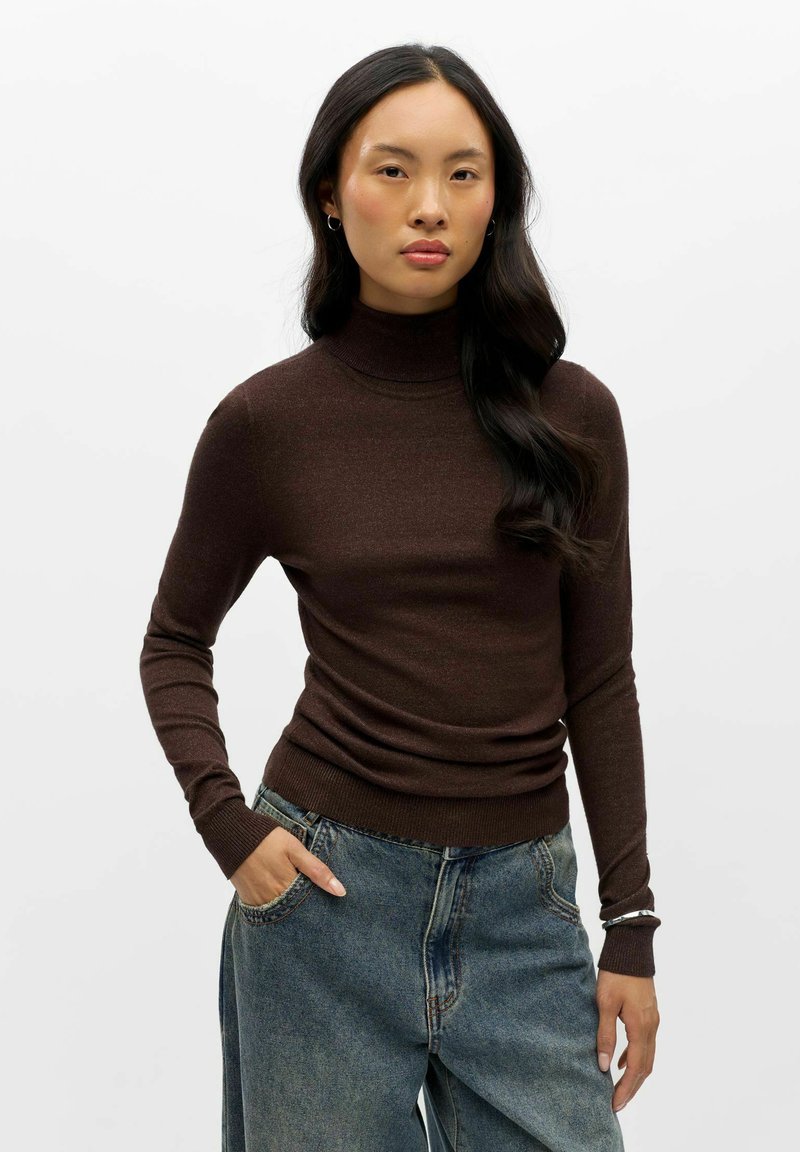 Femme aux longs cheveux foncés porte un pull à col roulé marron et un jean bleu taille haute, debout avec une main dans la poche contre un fond blanc.