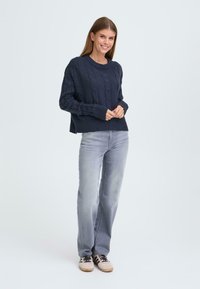 Marineblå kabelstrikket genser med ribbestrikkede mansjetter og nederkant, kombinert med lysegrå jeans og beige joggesko med svarte striper.