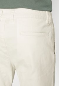 Pantalons en coton beige avec une texture lisse, présentant une poche arrière avec un subtil contour cousu. La ceinture est unie.
