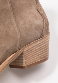 Stivaletto in suede marrone con tacco impilato a coste e dettagli cuciti. La vista ravvicinata mette in risalto la texture e i dettagli della forma.