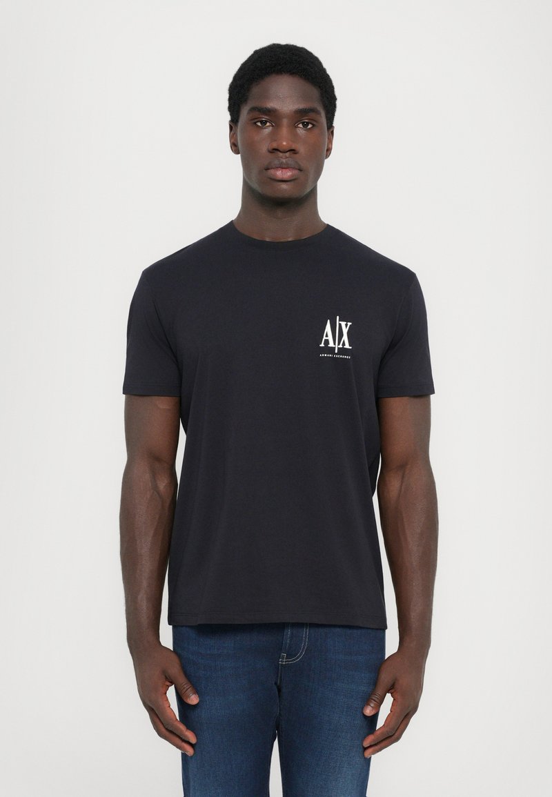 Svart t-skjorte med korte ermer og rund hals, med en hvit logo på venstre bryst. Kombinert med blå jeans.