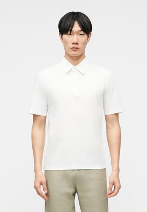 Un jeune homme se tient face à l'avant, portant un polo blanc uni à manches courtes et un pantalon beige clair, sur un fond uni.