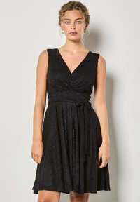 Czarna sukienka kopiująca z dekoltem w kształcie V, bez rękawów, z teksturowanej tkaniny i wiązanym detalem w talii, sięgająca powyżej kolana.