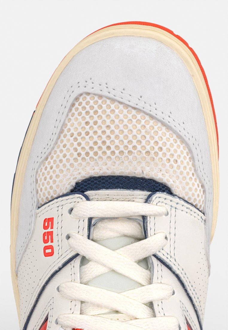 Weißer Sneaker mit Mesh-Einsätzen, einer Gummisohle und hellgrauen Wildlederelementen. Rote und blaue Akzente umfassen ein "550"-Logo an der Seite.