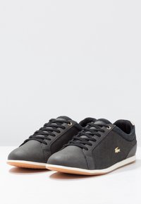 Ett par svarta låga sneakers med svarta snören, beige sulor och en liten gul logotyp på utsidan, visade mot en vit bakgrund.