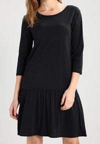 Robe noire mi-longue en tissu doux avec manches trois-quarts et ourlet froncé à volants. Encolure ronde, design minimaliste.