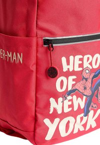 Röd tygryggsäck med ett spindeldesign, har en framficka med dragkedja och texten "HERO OF NEW YORK" i vitt. Utsmyckad med en rund spindelberlock.