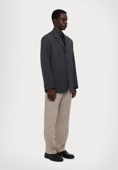 Hall kandiga klambriga ja kolme esiknöpiga hall bleiser, mis on ühendatud heledate beežide tekstuuriga pükstega ja mustade kingadega.