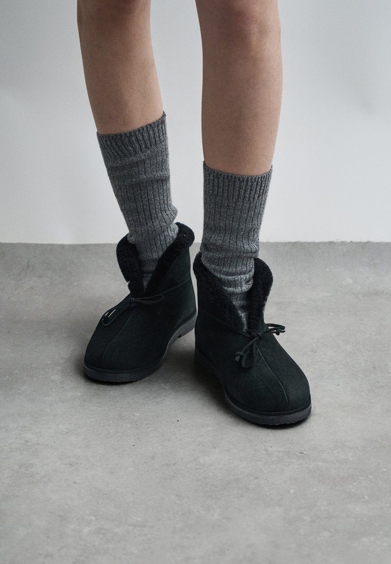 Bottes en daim noir avec doublure en fausse fourrure douce et liens ajustables, associées à des chaussettes gris côtelé, posées sur une surface en béton.