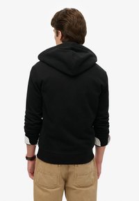 Schwarzer Hoodie mit großer Kapuze, gerippten Bündchen und Saum, getragen über einem weißen Hemd. Kombiniert mit hellbraunen Hosen mit Gesäßtaschen.