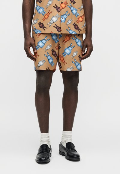 Homme portant un short et une chemise assortis couleur sable avec un motif de robots colorés, associés à des chaussettes blanches et des mocassins noirs, debout sur un fond uni.