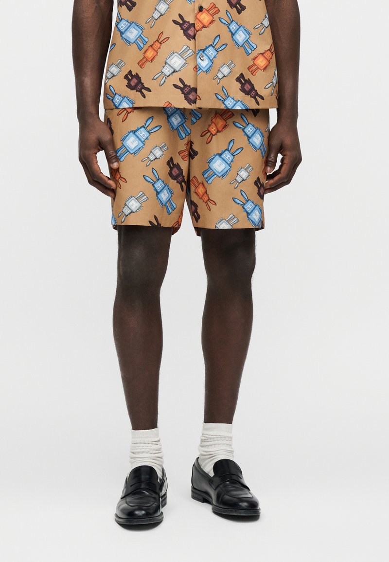 Άνδρας που φοράει ταιριαστά ταμπά  σορτς και πουκάμισο με πολύχρωμο σχέδιο ρομπότ, συνοδευόμενα από λευκές κάλτσες και μαύρα μοκασίνια, στέκεται σε απλό φόντο.