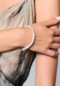 Perłowa bransoletka z naprzemiennie ułożonymi białymi perłami i złotymi akcentami, noszona na ręce na tle delikatnej, beżowo-niebieskiej tkaniny.