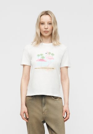 Fiatal nő fehér pólóban, amelyen rózsaszín flamingó, pálmafák és strandernyő látható, bézs nadrággal párosítva.