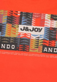 Heldere oranje t-shirt met een multicolore bladdessin en vetgedrukte tekst met "J&JOY" en "ANDO," gemaakt van zachte katoen.