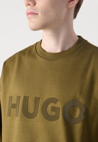 Felpa verde oliva con un grande logo lucido "HUGO" sul davanti, girocollo e polsini a costine. Tessuto dalla consistenza liscia.