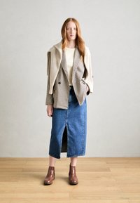 Blazer a motivi pied de poule beige, indossato su una maglietta crema. Gonna di jeans blu con spacco frontale e stivaletti marroni fino alla caviglia. Sfondo in legno.