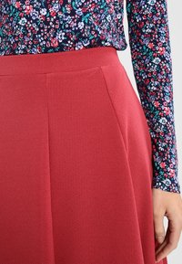 Falda roja texturizada con una cinturilla ajustada y pliegues en la parte delantera, combinada con una blusa de manga larga con un patrón floral sobre un fondo oscuro.