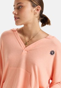 Licht koraal kleurige pullover met een V-hals, gemaakt van zachte stof, met een voorzak en een zon-embleem in marineblauw aan de linkerkant.