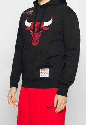 Person trägt schwarzen Hoodie mit rotem Chicago Bulls-Logo und rote Nike-Basketballshorts vor einem einfarbigen hellen Hintergrund.