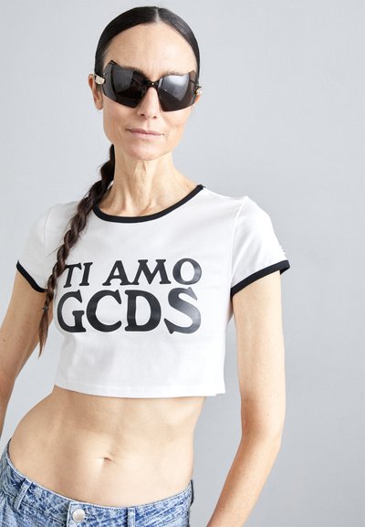 T-shirt branca cortada com bordas pretas e texto em negrito preto a dizer "TI AMO GCDS." O modelo usa óculos de sol pretos angulares e calções de ganga.