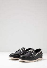 Zapatos de barco de cuero negro que cuentan con costuras blancas, agujetas blancas en contraste y una suela de goma beige con el emblema de la marca en el lateral.