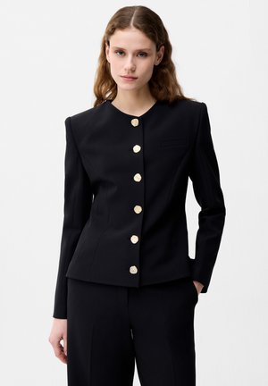 Kobieta ubrana w dopasowaną czarną marynarkę z złotymi guzikami oraz dopasowane czarne spodnie na jednolitym białym tle.
