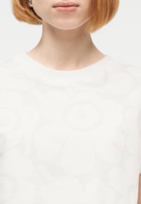 Άτομο με κοντά κόκκινα μαλλιά που φοράει λευκό τοπ με μεγάλα, διακριτικά λουλουδάτα σχέδια σε ανοιχτούς γκρι τόνους.