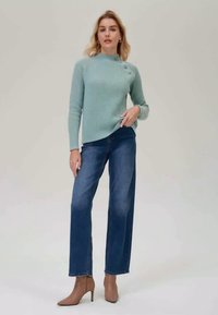 Helles blaues geripptes Sweatshirt mit einem hohen Kragen und drei Knöpfen auf einer Seite, kombiniert mit gerade geschnittenen Jeans und nudefarbenen High Heels.