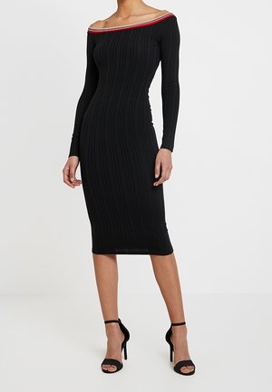 Vrouw draagt een aansluitende, lange mouwen, zwarte geribbelde midijurk met een off-shoulder halslijn met rode en witte biezen.