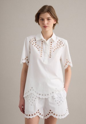 Femme portant une chemise blanche à manches courtes et un short assorti avec des bords festonnés et des motifs floraux découpés en forme circulaire.