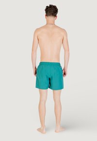 Pantaloncini da bagno teal con vita elasticizzata, lunghezza sopra il ginocchio e un piccolo logo sul lato sinistro. Tessuto liscio, senza fantasie.