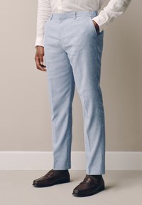 Man wearing light blue trousers, white long-sleeve shirt, dark brown loafers, standing with one hand in pocket against beige wall.