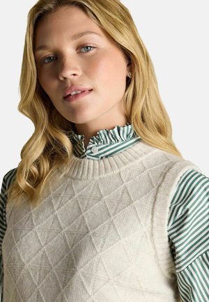 Blonde Frau mit blauen Augen trägt eine cremefarbene Strickweste über einer grün-weiß gestreiften Bluse mit Rüsche vor weißem Hintergrund.