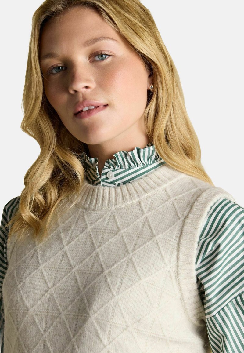 Blonde Frau mit blauen Augen trägt eine cremefarbene Strickweste über einer grün-weiß gestreiften Bluse mit Rüsche vor weißem Hintergrund.