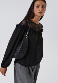Donna in blusa nera a maniche lunghe con lavorazione in pizzo e pantaloni grigi, che porta una borsa a spalla in pelle nera, mentre guarda di lato.