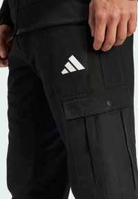 Black cargo pants with a smooth texture, featuring a large side pocket, snap closure, and a white Adidas logo on the thigh.