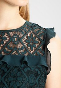 Blusa de encaje verde oscuro con intrincados patrones geométricos, acentos de hombros con volantes y un escote liso.