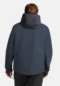 Giacca impermeabile di colore blu scuro con colletto alto e cappuccio, maniche lunghe e tasche laterali con zip. Presenta accenti a contrasto.