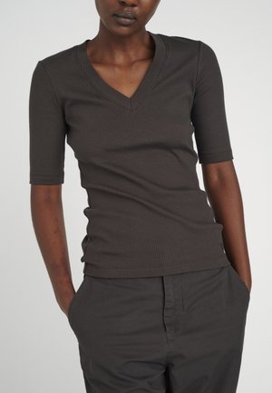 Vrouw draagt een getailleerd, geribd, donkergrijs V-hals shirt met ellebooglange mouwen en een donkergrijze broek, met haar handen in de zakken.