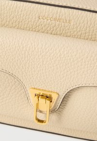 Béžová kožená kabelka s texturovaným povrchem, vybavená zlatou kovovou sponou a raženým logem. Zaoblený design s jemnými švy jako akcenty.