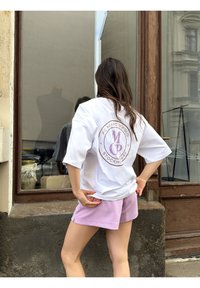 Γυναίκα με μακριά σκούρα μαλλιά στέκεται μπροστά σε ένα παράθυρο, φοράει ένα λευκό φαρδύ πουκάμισο με λογότυπο και ανοιχτόμωβ σορτς.