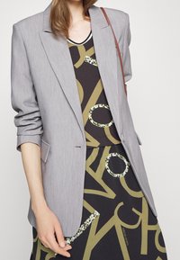 Hellgrauer, taillierter Blazer mit hochgeschobenen Ärmeln, kombiniert mit einem dunklen Kleid mit auffälligen grafischen Mustern in Oliven- und Grüntönen.