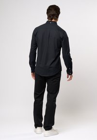 Schwarzes Langarmshirt mit einem knopfbaren Kragen und Manschetten, mit abgerundetem Saum und glatter Textur, kombiniert mit schwarzen Hosen und weißen Schuhen.