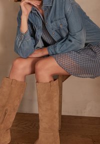 Personne assise sur un tabouret en bois, portant une robe à motifs, une veste en jean et des bottes en daim beige montant jusqu'aux genoux, reposant le menton sur la main.