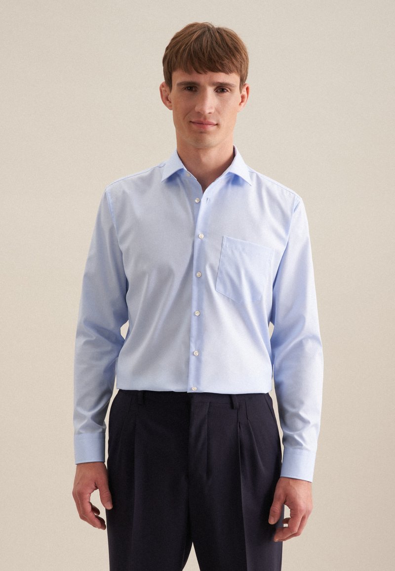 Chemise à manches longues bleu clair avec un col pointu, une poche frontale et des boutons blancs. Assortie avec un pantalon sombre, texture de tissu lisse.