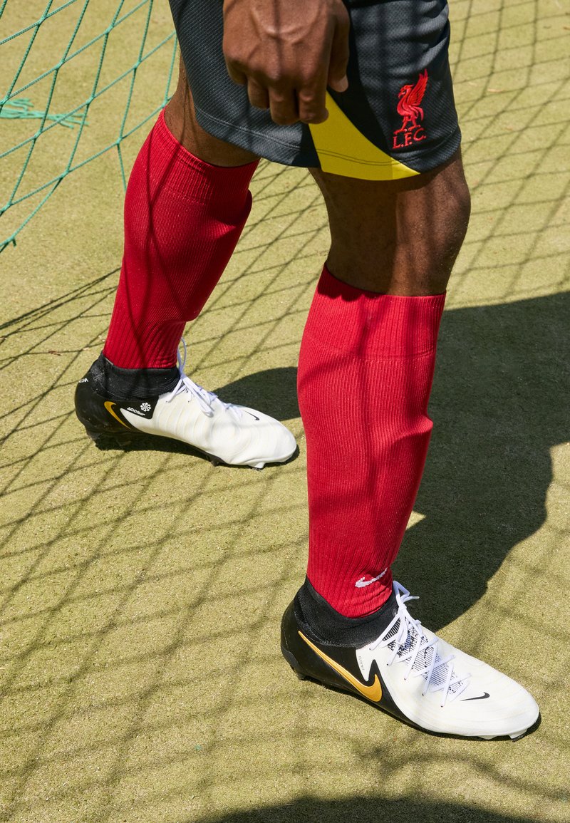 Scarpe da calcio bianche e nere con accenti dorati, parastinchi rossi e pantaloni grigi; in piedi su una superficie testurizzata con una rete verde sullo sfondo.