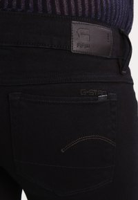 Pantalones de mezclilla negros con un parche de cuero, bolsillo trasero con costura curva y un acabado suave y texturizado.