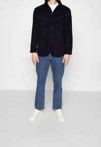Dark navy button-up shirt with two chest pockets, worn over a white t-shirt, paired with light blue flared denim jeans and white sneakers.